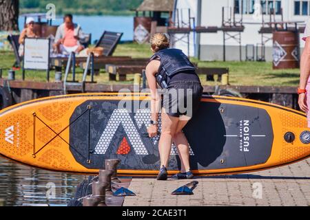 Steinhude, Germania, 29 maggio 2020: Giovane donna bionda in pantaloni corti neri trascina una tavola da surf sopra il molo nell'acqua del lago Foto Stock