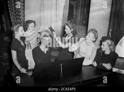 1945, storica, sei giovani donne attattive elegantemente vestite per una serata fuori in un salotto dell'hotel, divertendosi in piedi intorno al pianoforte dell'hotel che festeggia, con una delle giovani donne seduti al pianoforte che forse sta per suonare una canzone, USA. Foto Stock