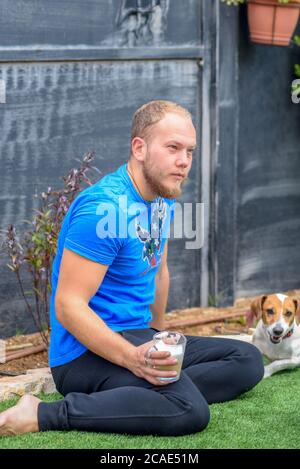 Il giovane atleta beveva caffè con latte da una tazza di vetro e il suo cane Jack Russell Terrier si siede accanto a lui e guarda nella macchina fotografica. Ritratto di un giovane uomo e cane outdoor. Foto Stock