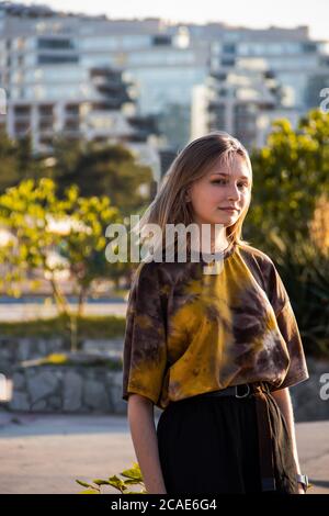 Ritratto di una giovane bella ragazza, bionda, in abiti alla moda, in piedi in un Parco sullo sfondo di alberi e edifici Foto Stock