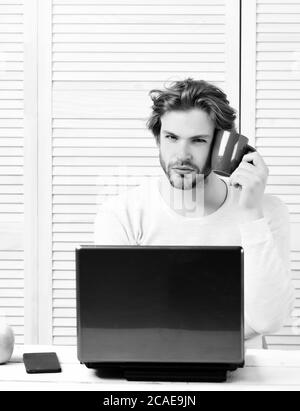 Freelance lavoro a casa e la pausa pranzo concetto. Uomo Barbuto con tazza vicino alla guancia sulla luce sfondo striato. Guy utilizza laptop e detiene un tè o un caffè mug. L'uomo con la faccia concentrata e gadget moderno Foto Stock