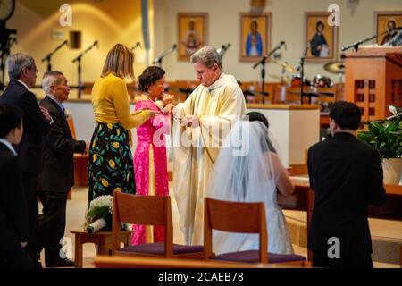 Il monsignor di una chiesa cattolica della California del sud offre la comunione agli ospiti in una cerimonia nuziale. Nota sposa e sposo a destra e musicista in Foto Stock