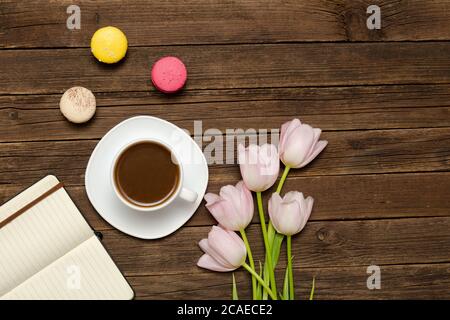 Tazza di caffè, macaron, tulipani rosa e taccuino su sfondo di legno. Vista dall'alto Foto Stock