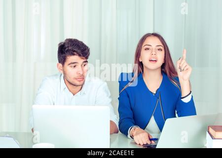 Giovane donna intelligente che tiene il dito in su avere un'idea luminosa mentre si siede con l'uomo che sta sbirciando spiando sul suo lavoro al tavolo nel soggiorno dell'ufficio a casa Foto Stock