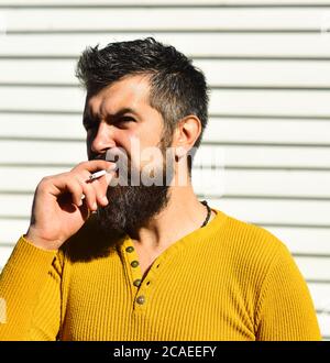 Cattiva abitudine e concetto di stile di vita malsano. Il ragazzo con la barba fuma all'esterno. Uomo con volto rigido su sfondo bianco a strisce. Il fumatore tiene la sigaretta in bocca. Foto Stock