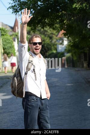 Studente con zaino in piedi fuori su sfondo stradale. Ragazzo in occhiali da sole onde e sorrisi. L'uomo con la barba e lo zaino sul retro gode di un viaggio. Concetto di viaggio e avventura Foto Stock