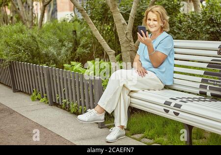 Donna bella bianca invecchiata circa 62 anni è seduta sulla panchina all'aperto con il telefono cellulare in mani durante il giorno in un panno casual. Sembra felice Foto Stock