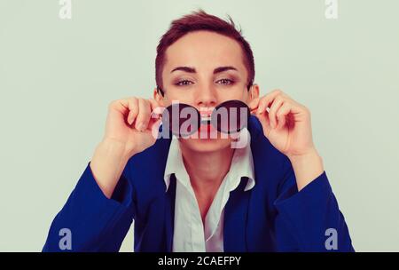 Ritratto di donna d'affari piuttosto giovane che guarda voi macchina fotografica che tiene gli occhiali da sole giù, che si pone in costume blu formale e bianco camicia a bottoni isolato Foto Stock