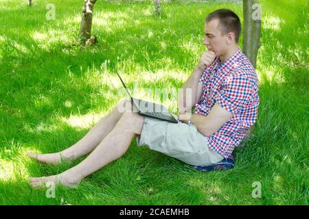 Il giovane si siede con un computer portatile nel giardino sull'erba. Lavori all'aperto Foto Stock