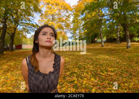 Giovane bella donna asiatica pensando e guardando in su circondato da alberi d'autunno scenici e foglie nella foresta Foto Stock