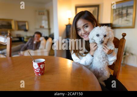 Giovane donna asiatica felice sorridente mentre tiene il cane carino all'interno della casa comoda Foto Stock