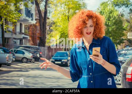 Chiamare un taxi da app. Donna che mantiene il telefono cellulare e il sollevatore escursionismo nella strada di città in una giornata di sole. Una sola persona. Il segnale di arresto. Stile di vita. Foto Stock