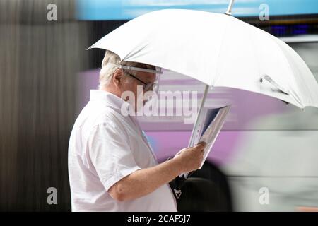 Belgrado, Serbia - 5 agosto 2020: Anziano uomo in camicia bianca che indossa uno scudo protettivo che cammina sotto l'ombrello mentre si guarda carte in plastica Foto Stock