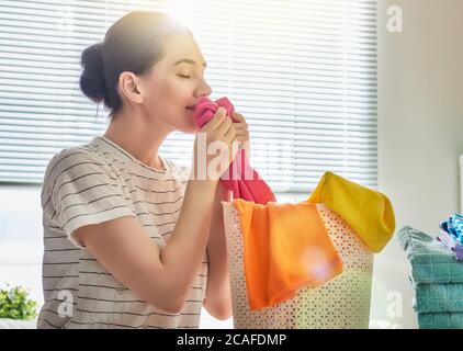 Bella giovane donna sta odorando vestiti puliti e sorridendo mentre facendo lavanderia a casa. Foto Stock