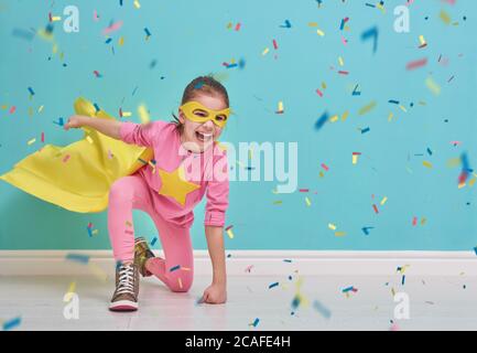 Il bambino piccolo gioca il supereroe. Capretto sullo sfondo della parete blu brillante. La ragazza sta gettando i confetti. Colori giallo, rosa e turchese. Foto Stock