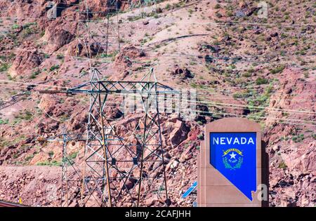 Benvenuto al cartello del monumento del Nevada. Linee di trasmissione di potenza ad alta tensione e torri sfocate che trasmettono elettricità da Hoover Dam. Terre rocciose del deserto Foto Stock