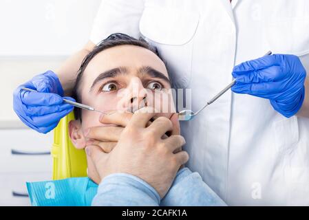 Uomo caucasico seduto sulla sedia del dentista, le sue mani che ricoprono la bocca, spaventato e preso dal panico Foto Stock