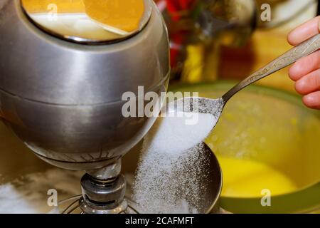 Le mani femminili aggiungono uno zucchero alla ciotola del miscelatore con gli albumi a montare per fare una torta. Foto Stock