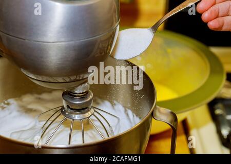 Il mixer che monta gli albumi per la torta. Donna aggiunge zucchero al recipiente del miscelatore. Foto Stock