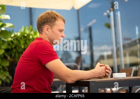 Uomo serio biondo in stile casual t-shirt rossa sedersi in strada moderna caffetteria, nervoso attesa qualcuno che guarda su sedia vuota. Relazione umana Foto Stock