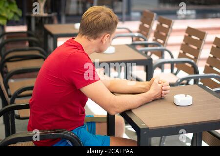 Uomo serio biondo in stile casual t-shirt rossa sedersi in strada moderna caffetteria, nervoso attesa qualcuno che guarda su sedia vuota. Relazione umana Foto Stock