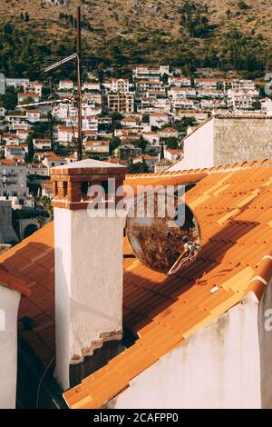 Primo piano di un camino e antenne di piatto sulle tegole arancioni del tetto di una casa contro lo sfondo di edifici residenziali e vista sugli alberi. Foto Stock