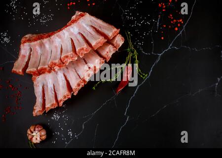 Costolette di maiale crude con erbe, aglio e peperoncino rosso su fondo nero. Foto Stock