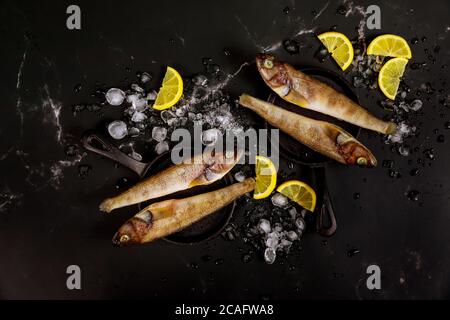 Pesce intero crudo con ghiaccio e limone su tavola nera. Vista dall'alto. Foto Stock