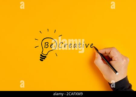 La mano maschile scrive le parole essere creativi e disegna una lampadina su sfondo giallo. Creatività e innovazione. Foto Stock