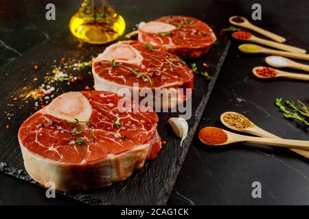 Gambo di manzo crudo in osso e spezie per la preparazione di piatto di ossobuco su fondo nero. Foto Stock