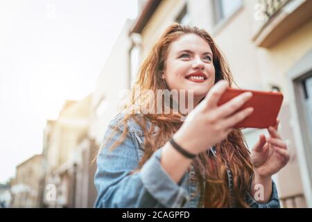 Sorridente rosso ricci lunghi capelli caucasici teen girl camminando per la strada e navigando in internet utilizzando il moderno smartphone. Persone moderne con tecnologia Foto Stock
