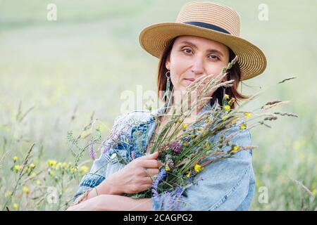 Ritratto di una giovane donna calma con bouquet di fiori selvatici. Vestì una giacca di jeans, un cappello di paglia e un abito estivo leggero. La bellezza delle persone naturali nel Foto Stock