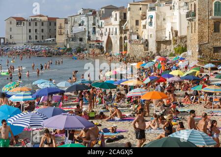 11.08.2018, Cefalu, Sicilia, Italia - turisti e gente del posto si rilassano sulla spiaggia della città sotto gli ombrelloni e si bagnano nel Mar Mediterraneo. Sullo sfondo Foto Stock