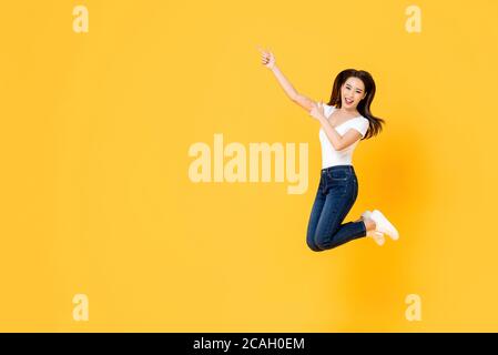 Felice sorridente giovane ragazza asiatica che salta con le mani che si accalcano isolato su sfondo giallo dello studio con spazio di copia Foto Stock