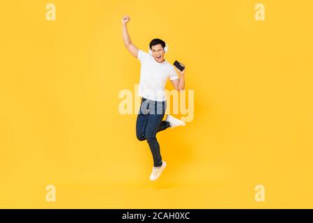Felice giovane asiatico che indossa le cuffie per ascoltare la musica telefono cellulare e salti con pugno di sollevamento isolato su giallo sfondo Foto Stock