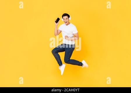 Felice giovane asiatico che indossa le cuffie per ascoltare la musica telefono cellulare e salti isolati su sfondo giallo Foto Stock