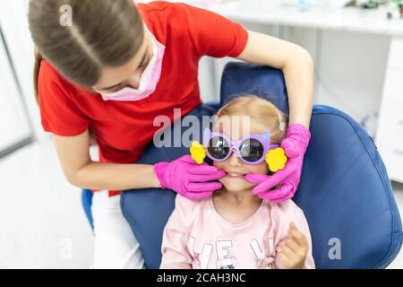 Bella ragazza giovane seduta tranquillamente nella sedia dei dentisti mentre guardando una dentista femmina che tiene gli strumenti dentali Foto Stock