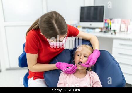Bella ragazza giovane seduta tranquillamente nella sedia dei dentisti mentre guardando una dentista femmina che tiene gli strumenti dentali Foto Stock