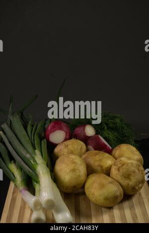 Patate giovani, ravanello, cipolle verdi, aneto su tavola di legno, fondo nero Foto Stock