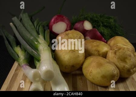 Patate giovani, ravanello, cipolle verdi, aneto su tavola di legno, fondo nero Foto Stock