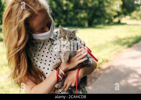 Giovane bionda in una maschera medica protettiva in una passeggiata con un gattino scozzese dritto in estate durante una pandemia di infezione da coronavirus covid 19. Foto di alta qualità Foto Stock