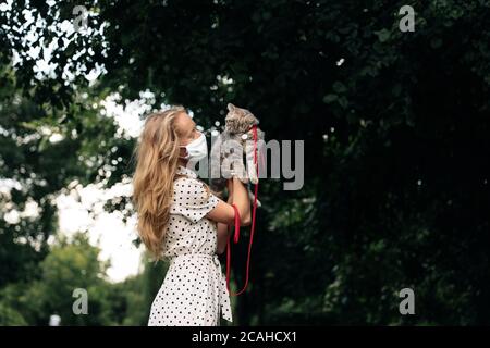 Una giovane bionda in una maschera medica protettiva tiene un gattino scozzese ad orecchie dritte. Una ragazza cammina con un gatto durante il coronavirus kovid19 pandemia con misure di sicurezza Foto Stock