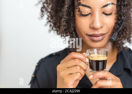 tazza trasparente con caffè espresso nero cremoso trattenuta da una bella donna afro-nera con cuffia per helpdesk. messa a fuoco selettiva sulla tazza Foto Stock