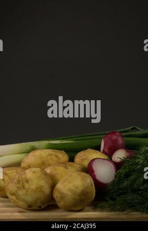 Patate giovani, ravanello, cipolle verdi, aneto su tavola di legno, fondo nero Foto Stock