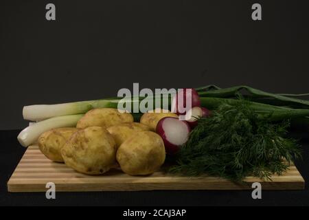 Patate giovani, ravanello, cipolle verdi, aneto su tavola di legno, fondo nero Foto Stock