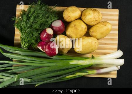 Patate giovani, ravanello, cipolle verdi, aneto su tavola di legno, fondo nero Foto Stock