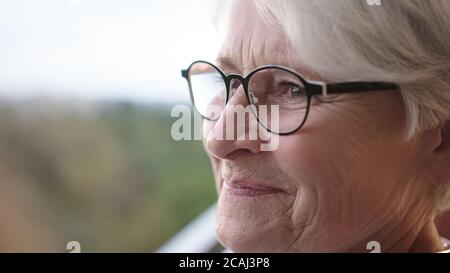 Donna anziana con capelli grigi e bicchieri in piedi da soli sul balcone con occhi sorridenti ma tristi. Filmati 4k di alta qualità Foto Stock