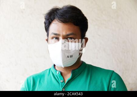 Uomo indiano adulto che indossa un respiratore bianco N95 in una T-Shirt verde rivolta verso la fotocamera e guardando verso di sé (occhi neri, maschera N95, sfondo bianco, luce naturale, Foto Stock