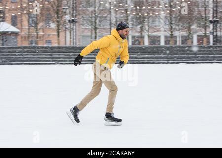 Ritratto di uomo bearded in giacca gialla, pantaloni beige, cappello nero su pista di ghiaccio, all'aperto nella giornata invernale innevata, vista laterale. Bell'uomo che spara in mot Foto Stock