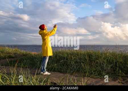 Uomo in giallo impermeabile indossare cappello rosso in piedi sulla spiaggia in legno percorso, guarda drammatico cielo nuvoloso e scatta foto su smartphone. Stagione autunnale. Foto Stock